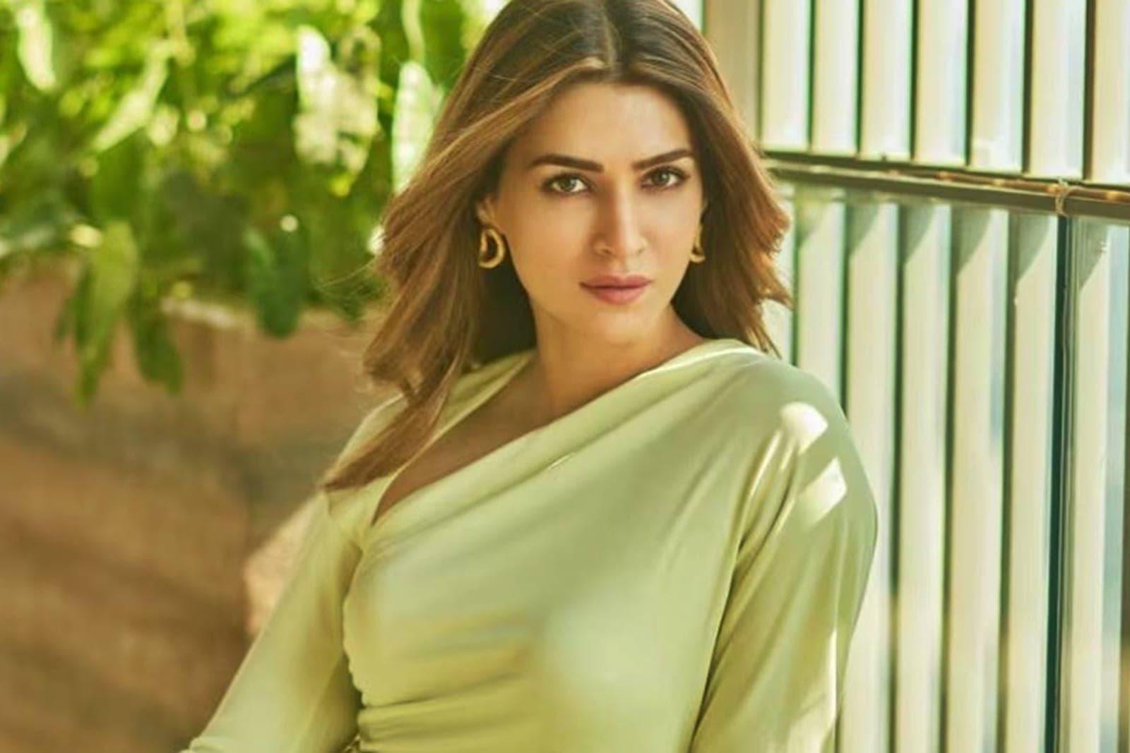 Kriti Sanon, a Bollywood actress, posing in a light lime green asymmetrical top with gold hoop earrings, looking directly at the camera with a serene expression. Her brown hair is wavy, and the background is blurred green foliage and modern architecture.