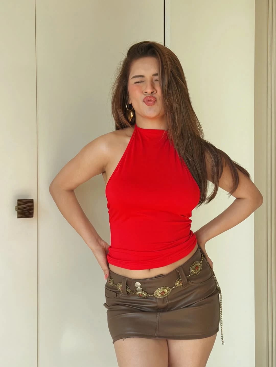 Avneet Kaur posing playfully with puckered lips and closed eyes, wearing a red halter top and a brown mini skirt with a decorative gold belt.