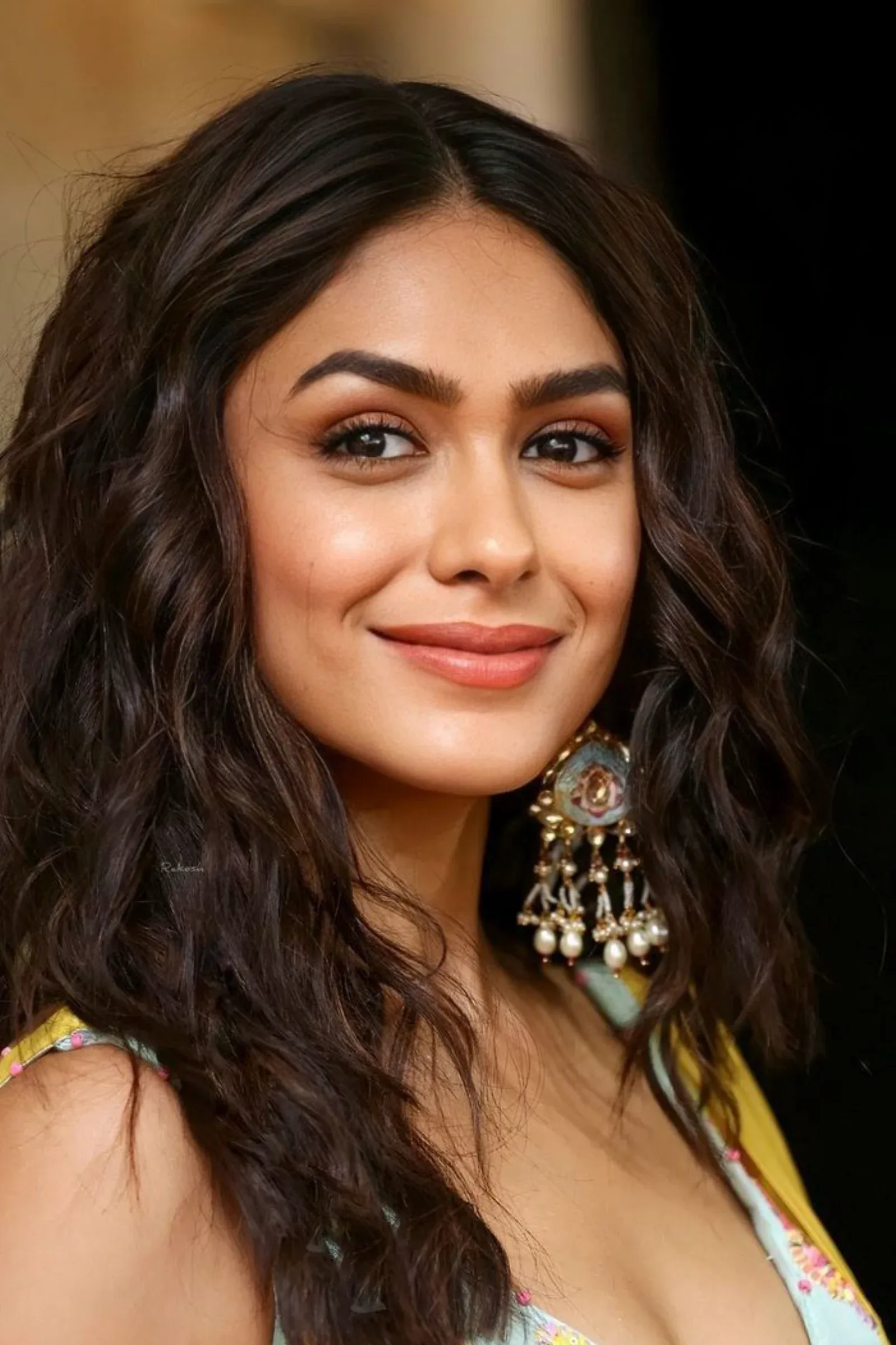 Close-up portrait of Mrunal Thakur smiling, with wavy dark hair and ornate traditional earrings.