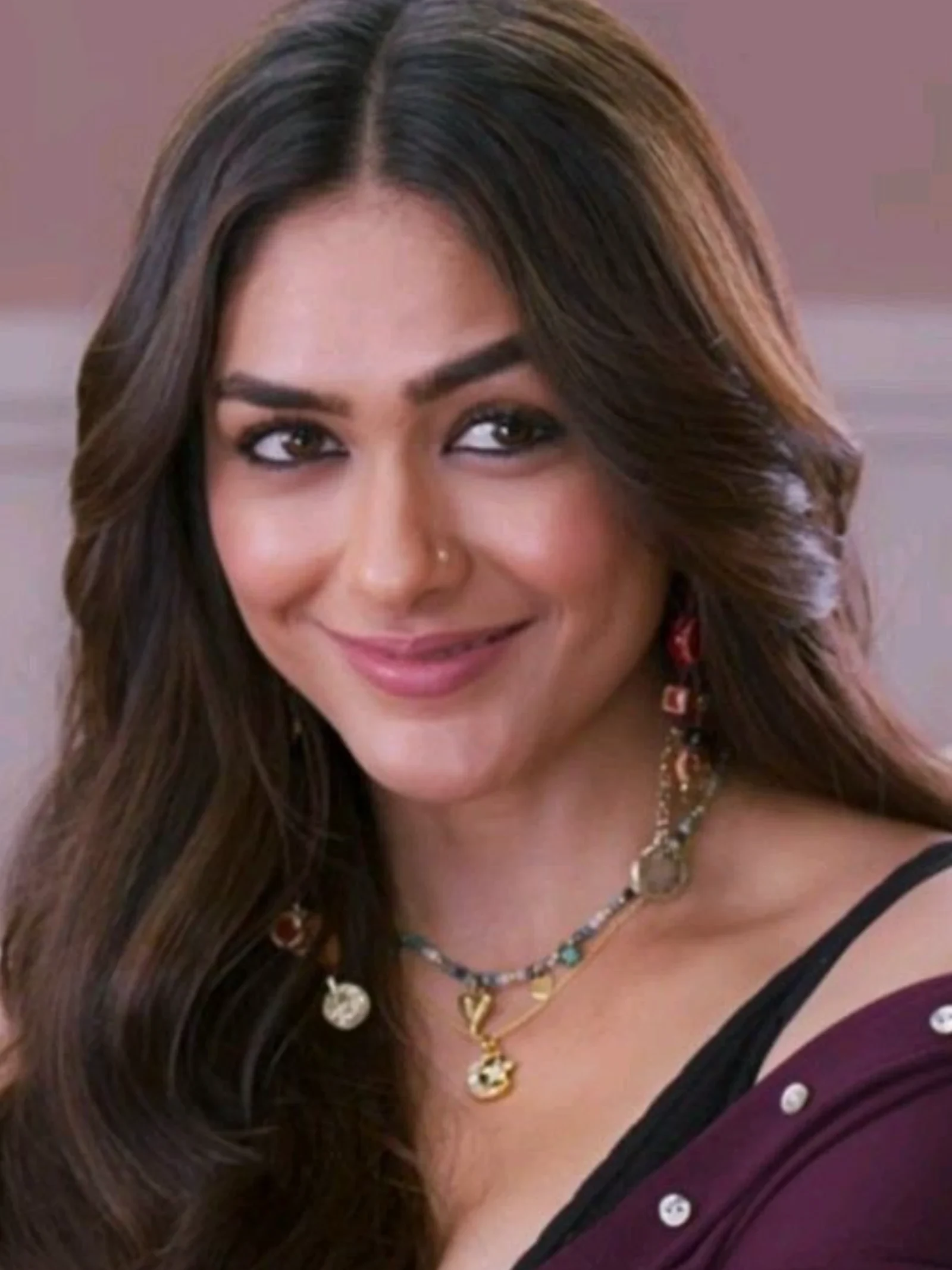 Close-up portrait of Mrunal Thakur smiling, wearing layered charm necklaces, multi-colored dangle earrings, and a dark plum top.