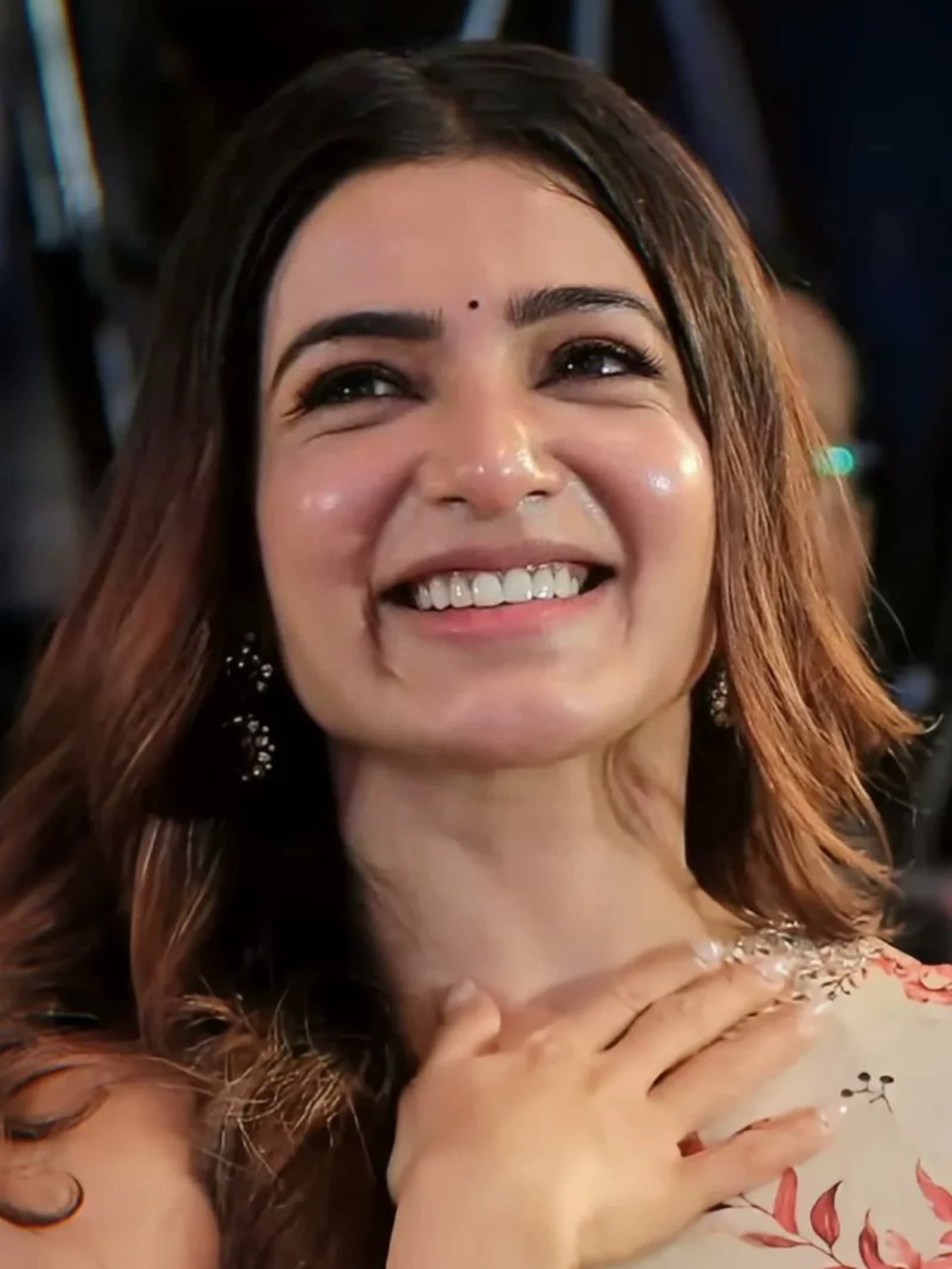 Close-up portrait of Samantha Ruth Prabhu smiling broadly, wearing a light-colored floral saree with red and pink patterns, a black bindi, and beaded earrings, with her hand on her chest.