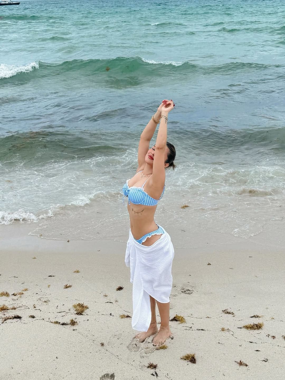 Avneet Kaur on a sandy beach, wearing a blue and white striped bikini and white sarong, stretching her arms overhead with a joyful smile, ocean waves in the background.
