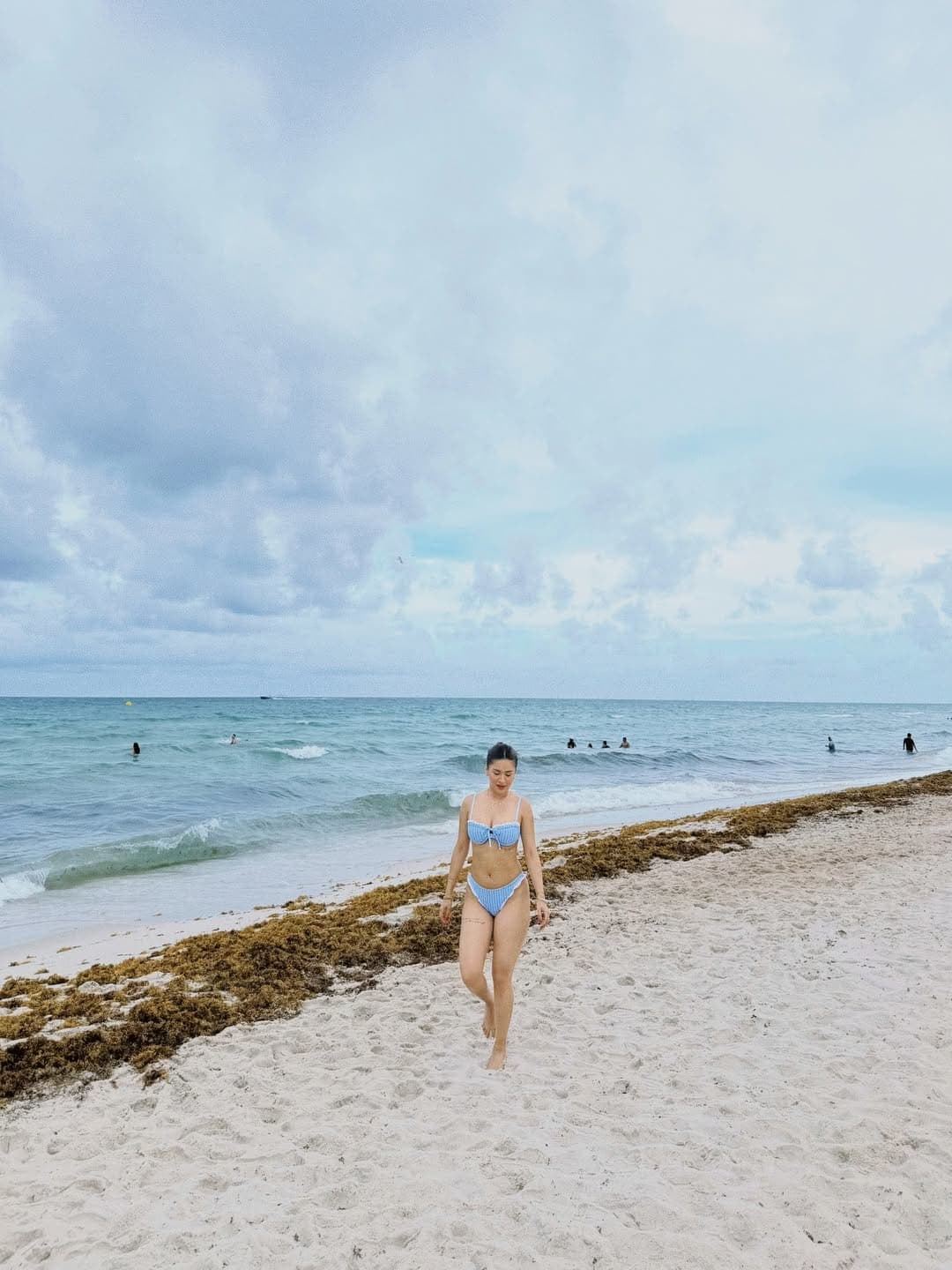 Avneet Kaur walking barefoot on a sandy beach, wearing a light blue and white striped bikini, with the ocean and people swimming in the background under an overcast sky.