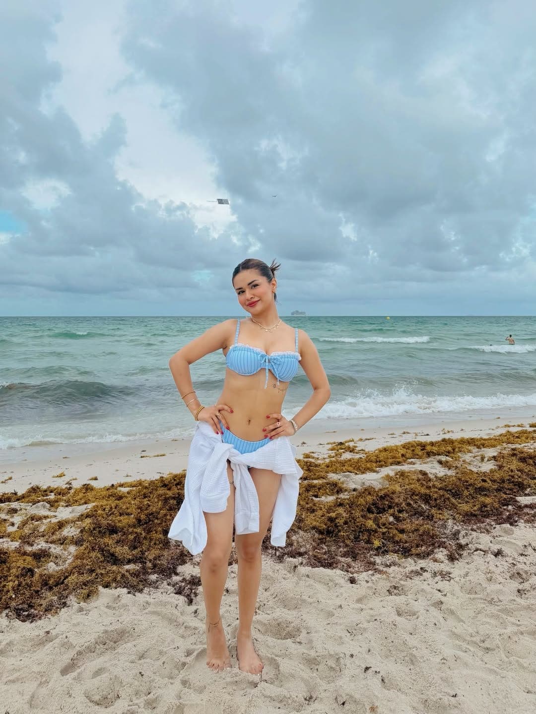 Avneet Kaur standing on a beach in a light blue striped bikini with a white shirt tied around her waist, hands on hips, looking confidently at the camera. The ocean and cloudy sky are in the background.
