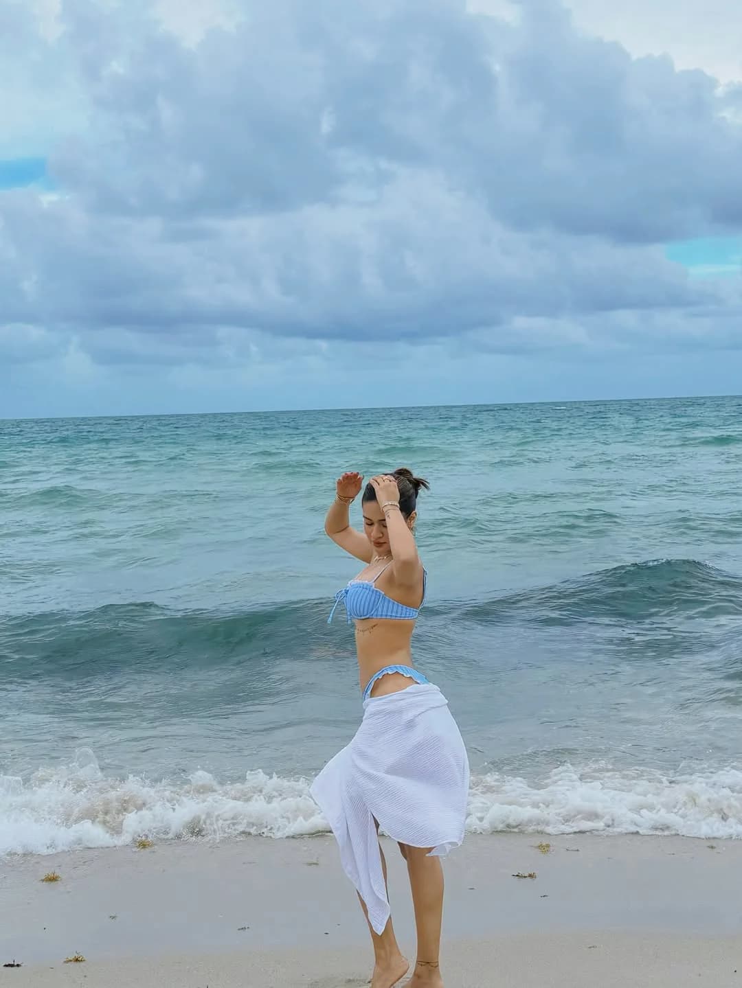 Avneet Kaur on a sandy beach, wearing a blue and white striped bikini top and a white sarong, with her hands raised, posing playfully by the ocean under a cloudy sky.