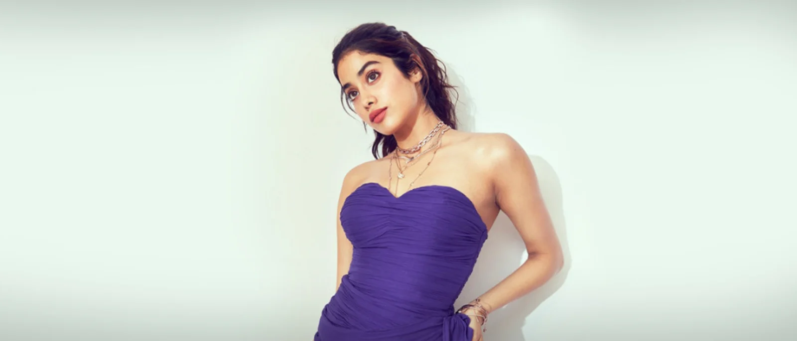 Janhvi Kapoor in a purple strapless ruched dress, looking up with layered necklaces against a light background.