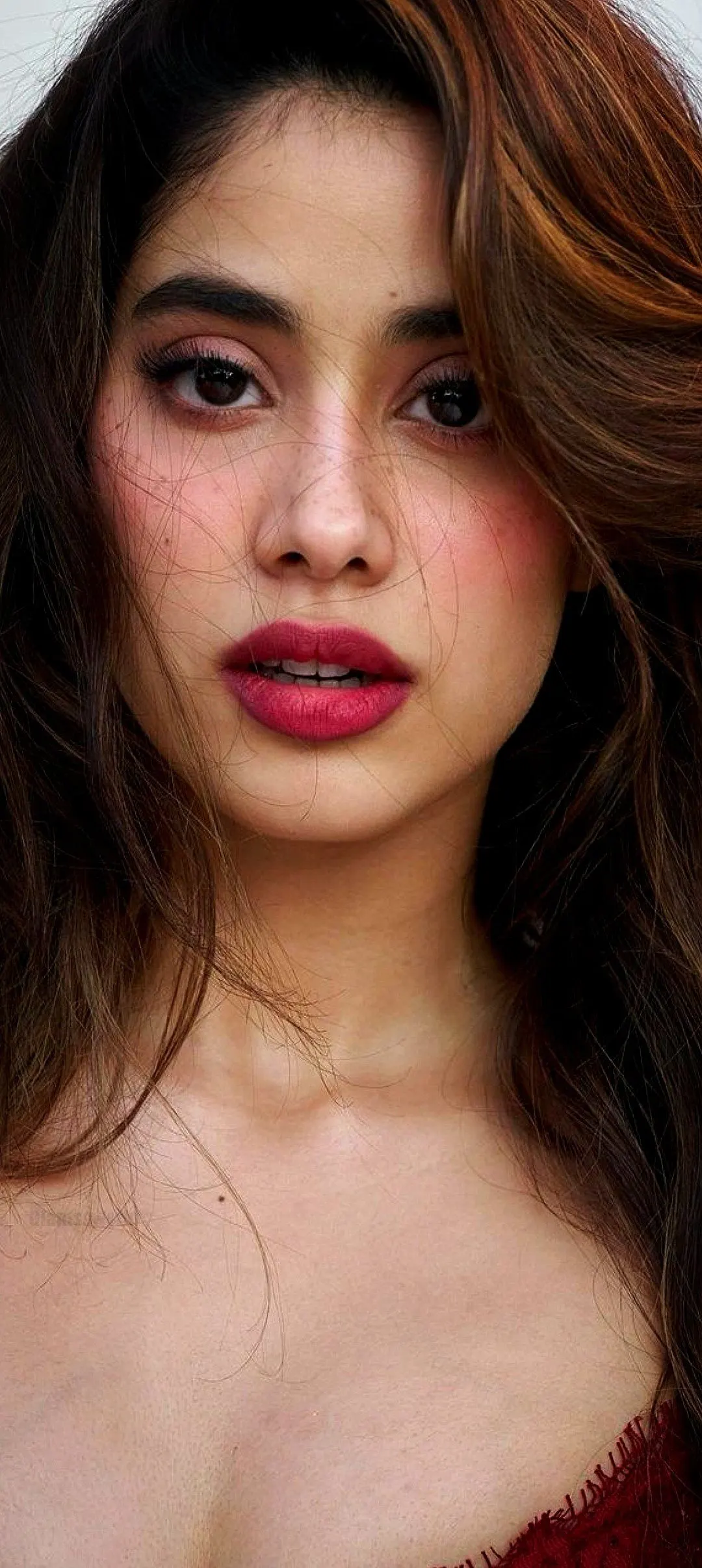 Close-up portrait of Janhvi Kapoor, an Indian actress, with long wavy brown hair, vibrant fuchsia lipstick, and expressive dark eyes, looking directly at the camera.