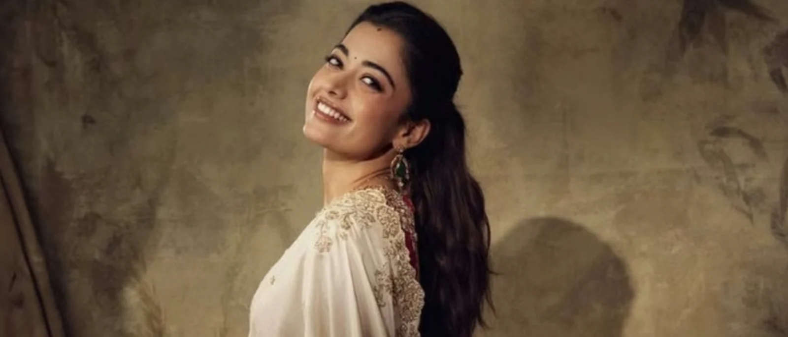 Rashmika Mandanna smiling brightly, looking over her shoulder, wearing an off-white traditional Indian outfit with gold embroidery and green drop earrings against a textured brown wall.