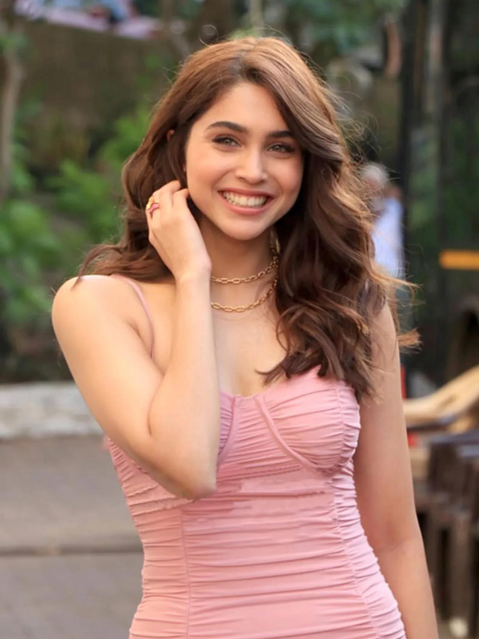 Sharvari Wagh smiling brightly, wearing a light pink ruched spaghetti strap dress and a gold chain necklace, with wavy brown hair.