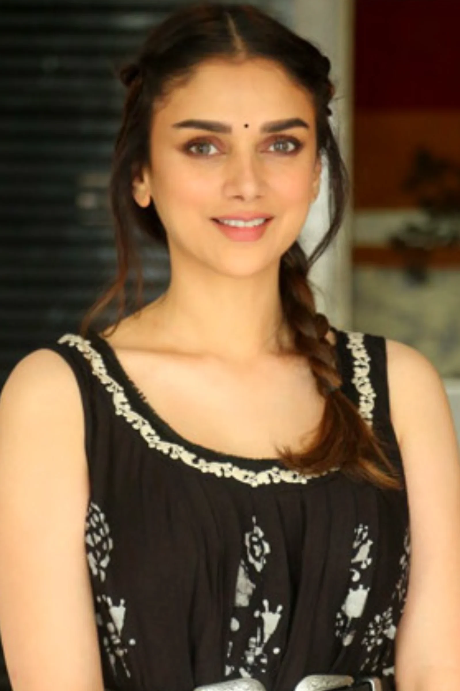 Aditi Rao Hydari smiling, wearing a black sleeveless dress with white patterns and a side braid, with a bindi on her forehead.