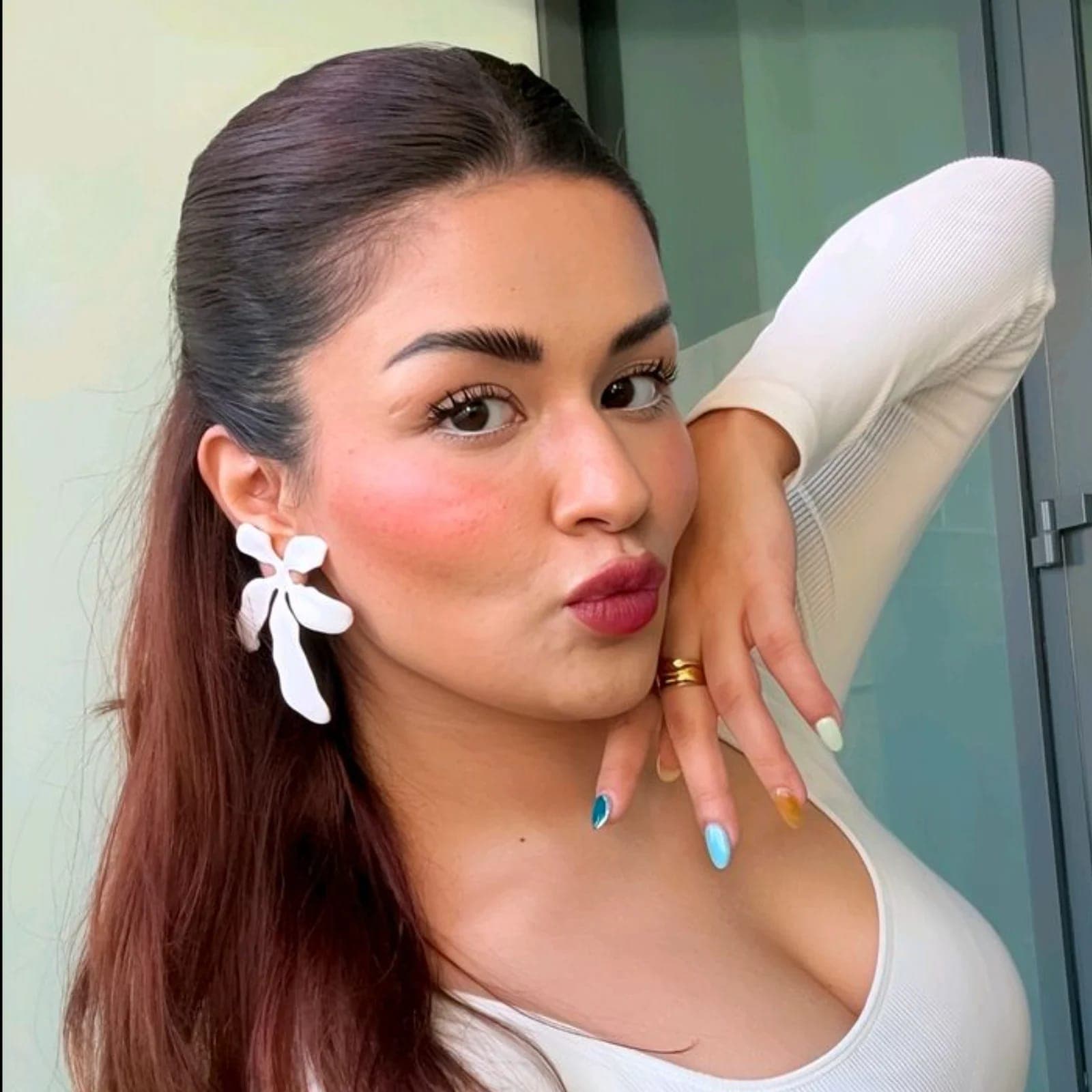 Close-up portrait of Avneet Kaur with dark brown hair, wearing a white ribbed top, large white floral earrings, and a gold ring. She has rosy blush, white eyeliner, berry lipstick, and multicolored nails, striking a playful pout.