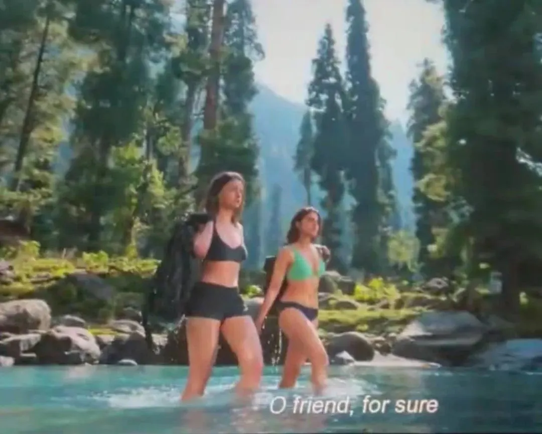 Alia Bhatt in a black bikini and Sharvari Wagh in a green bikini wading through a clear mountain stream in a lush forest.