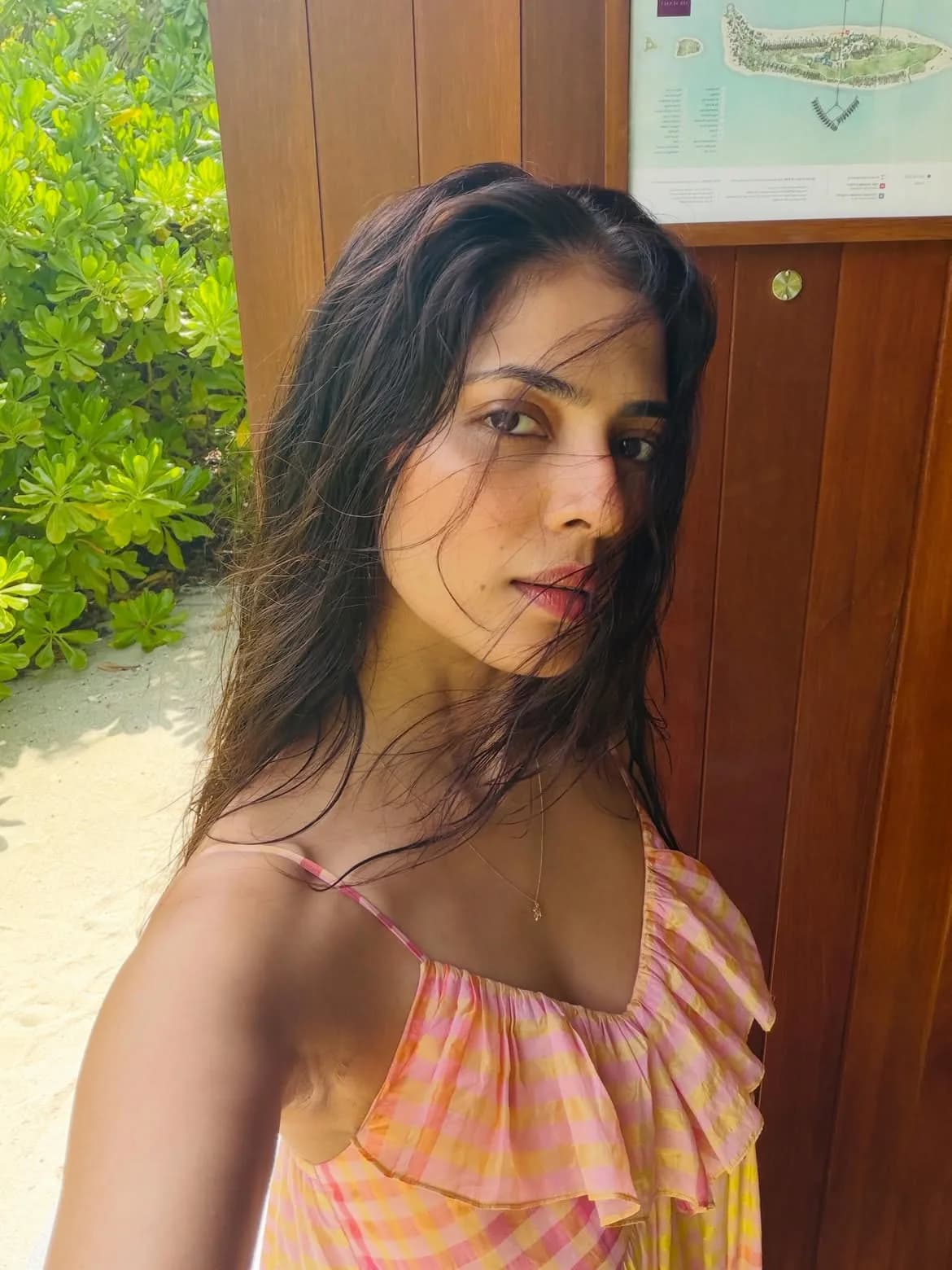 Malavika Mohanan taking a selfie in a pink and yellow plaid ruffled dress, with wet, dark hair, against a background of green tropical foliage and a wooden wall with a resort map.