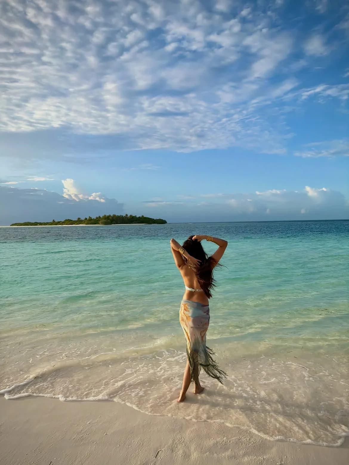 Malavika Mohanan from behind, standing on a white sand beach, looking at the turquoise ocean and a distant island under a blue sky. She wears a bikini top and a patterned sarong, with hands behind her head.