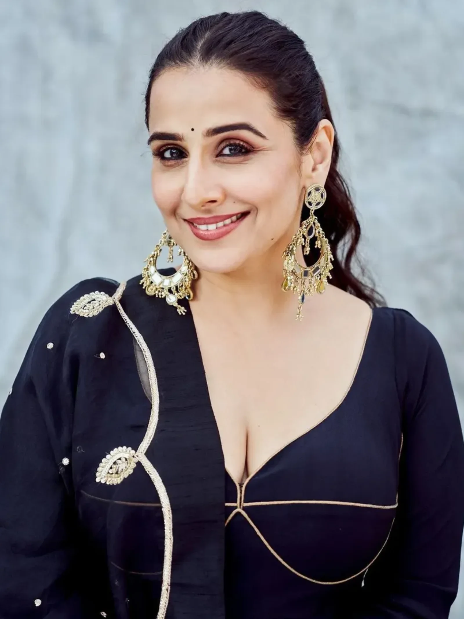 Vidya Balan smiling, wearing a black deep-neck blouse with gold piping and large gold chandelier earrings, against a grey background.