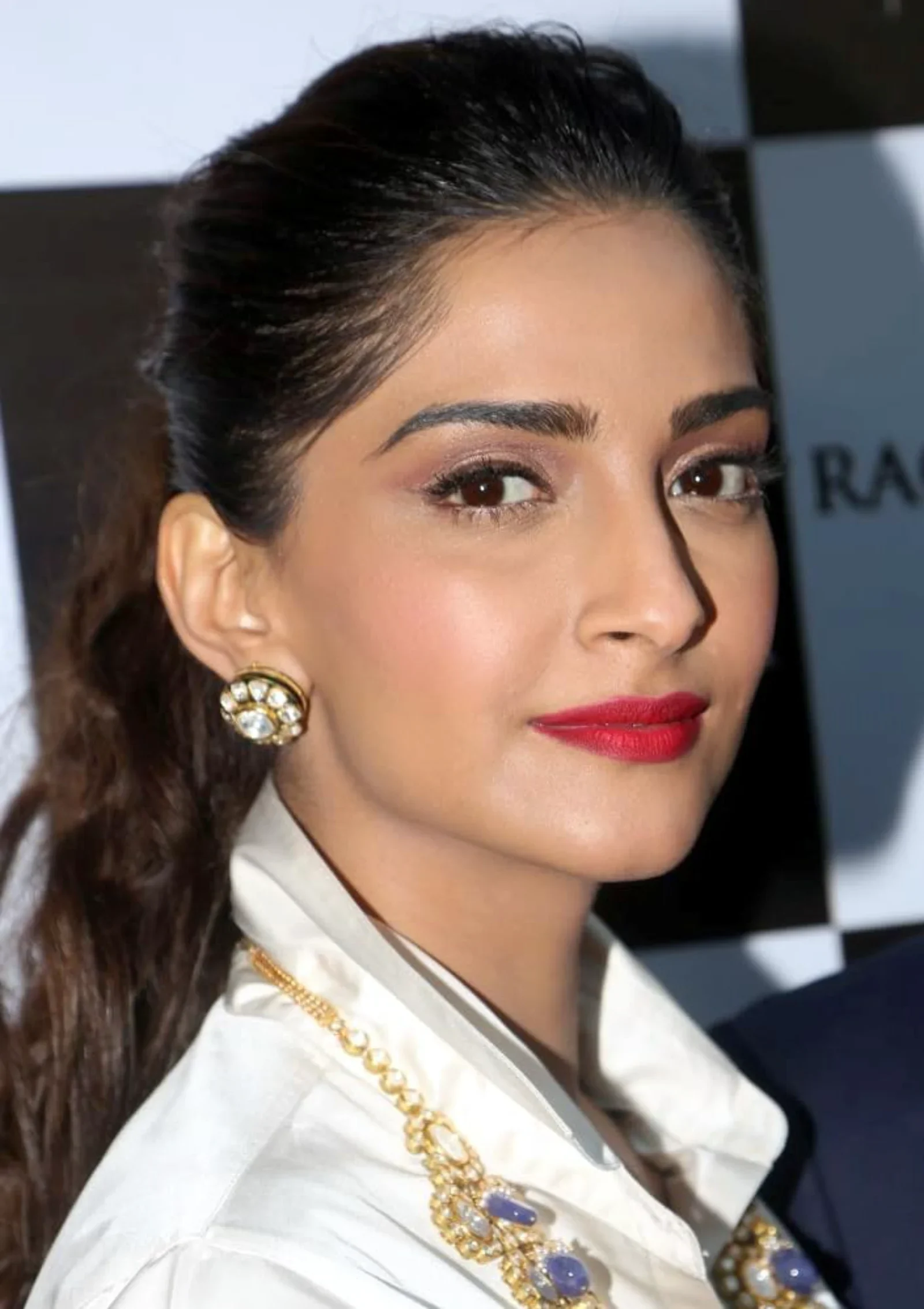 Close-up portrait of Sonam Kapoor wearing a white collared shirt, a gold necklace with purple gemstones, and matching earrings. She has dark hair pulled back, bold red lipstick, and a confident expression.