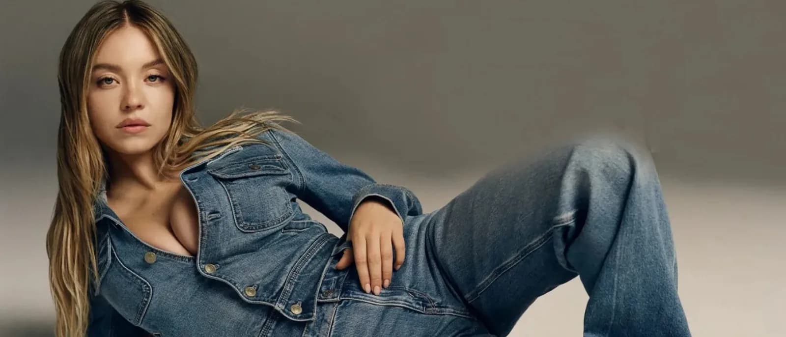 Sydney Sweeney reclining, wearing an open denim jacket and jeans, looking confidently at the camera.