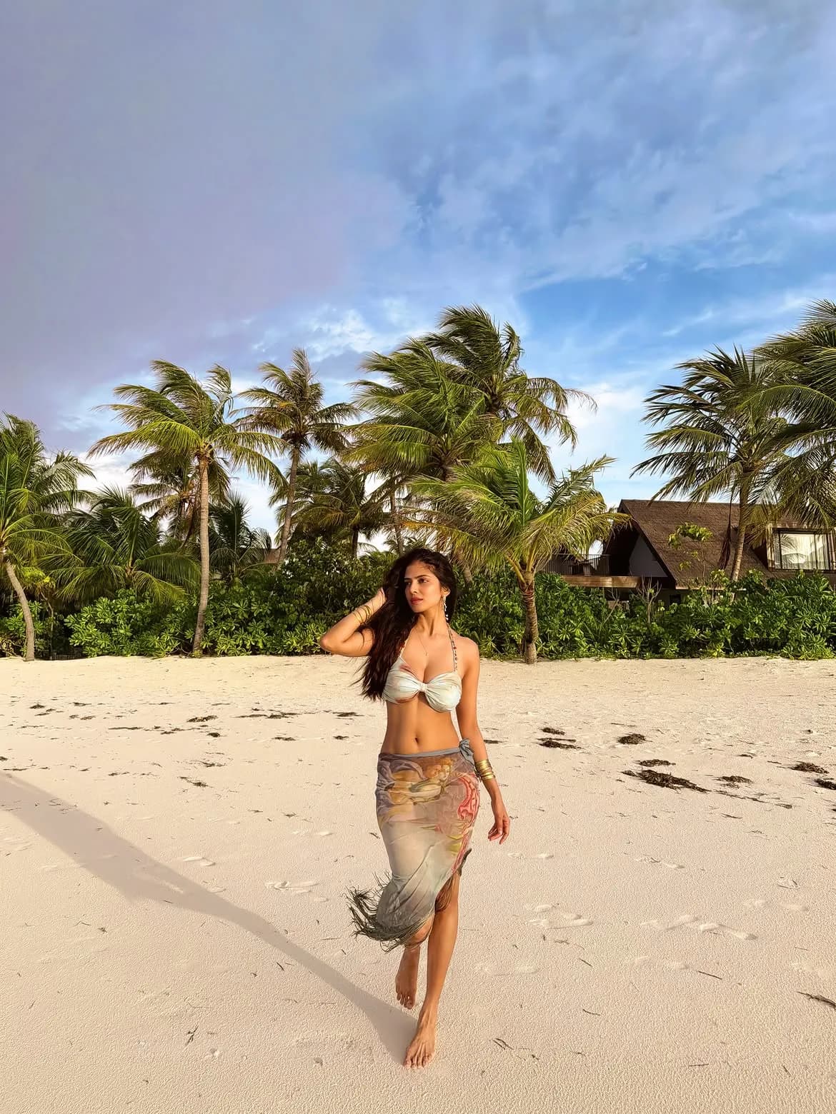 Malavika Mohanan walking barefoot on a white sand beach, wearing a light bikini top and a printed sarong, with palm trees and a resort in the background under a blue sky.