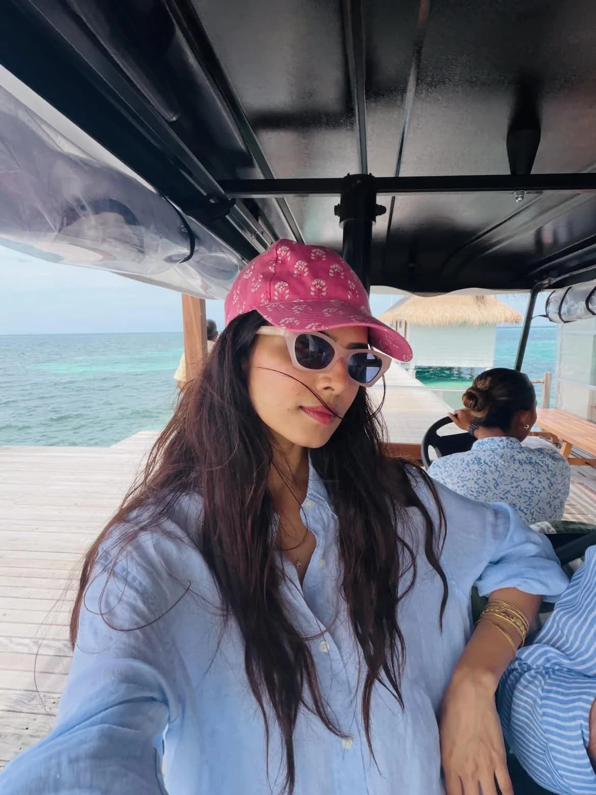 Malavika Mohanan wearing a pink patterned baseball cap, white cat-eye sunglasses, and a light blue linen shirt, sitting in a vehicle on a wooden pier over clear turquoise water in the Maldives.