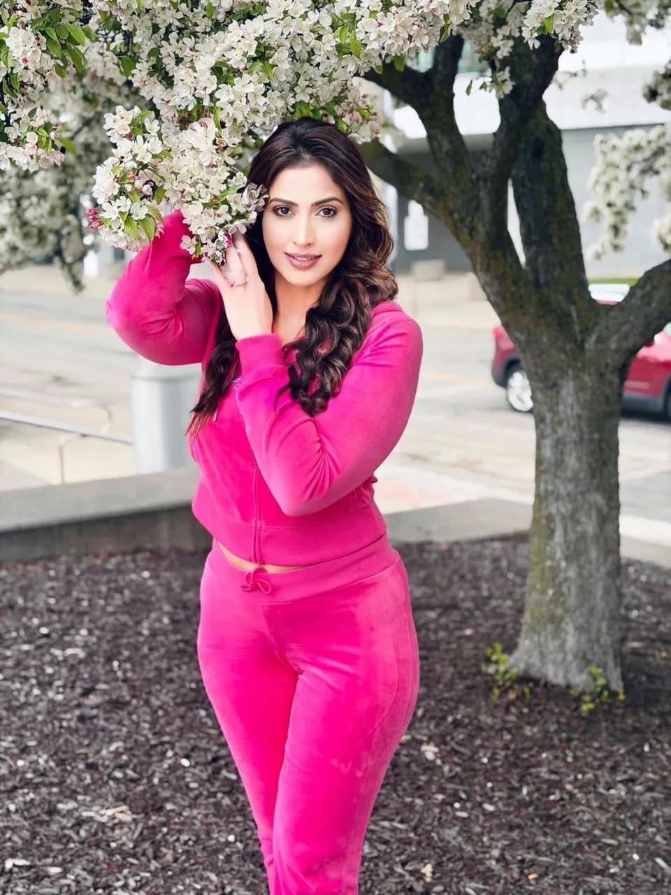Samaira Sandhu in a bright pink velour tracksuit, smiling and posing under a tree with white blossoms.