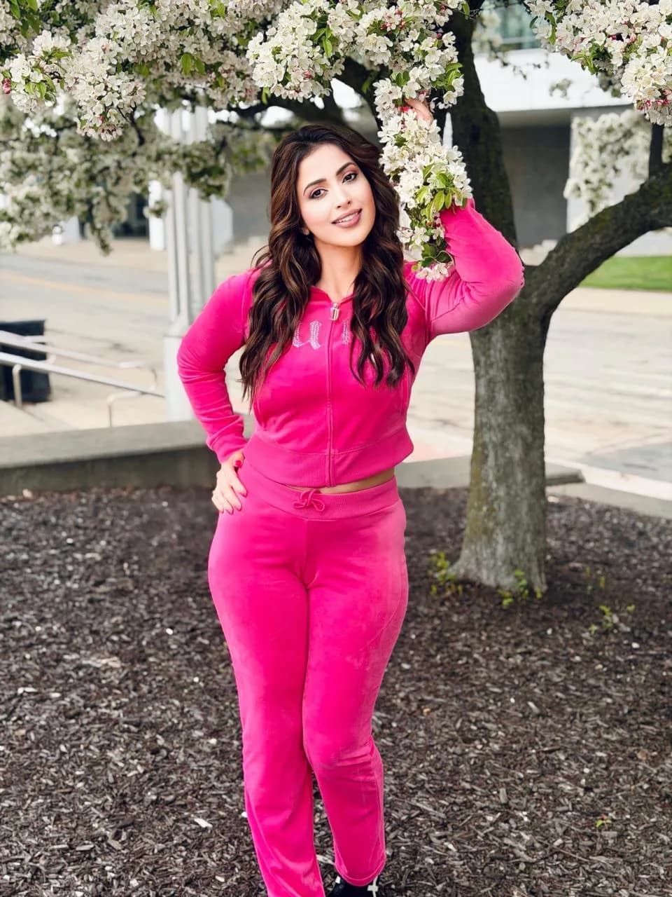 Samaira Sandhu smiling, wearing a bright hot pink velour tracksuit, standing under a tree with white blossoms, with one hand on her hip and the other touching a branch.