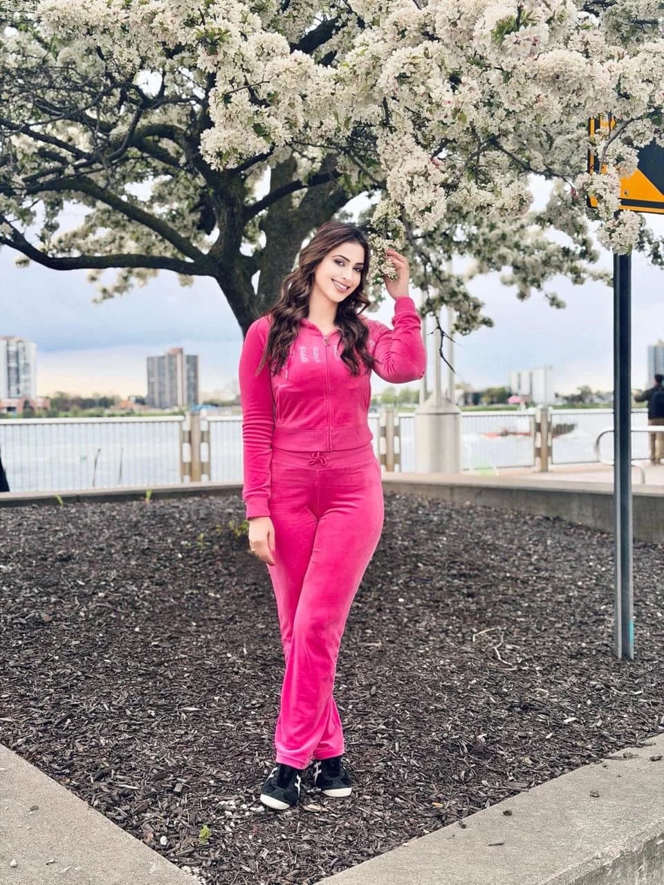 Samaira Sandhu smiling, wearing a hot pink velvet tracksuit, standing under a tree with white blossoms, with a waterfront and city buildings in the background.