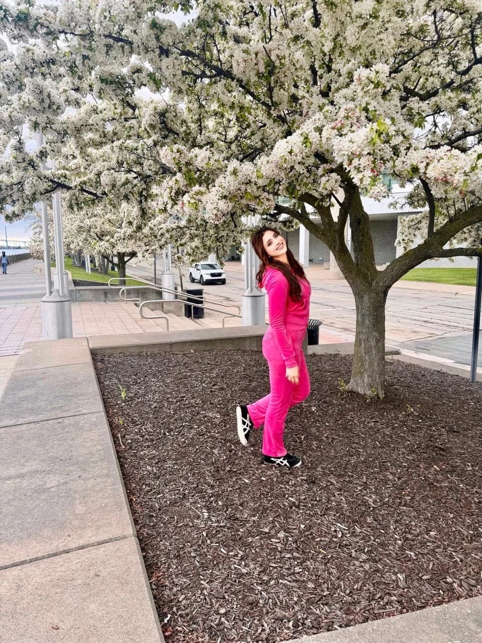 Samaira Sandhu smiling brightly in a hot pink velour tracksuit under a tree with white blossoms.