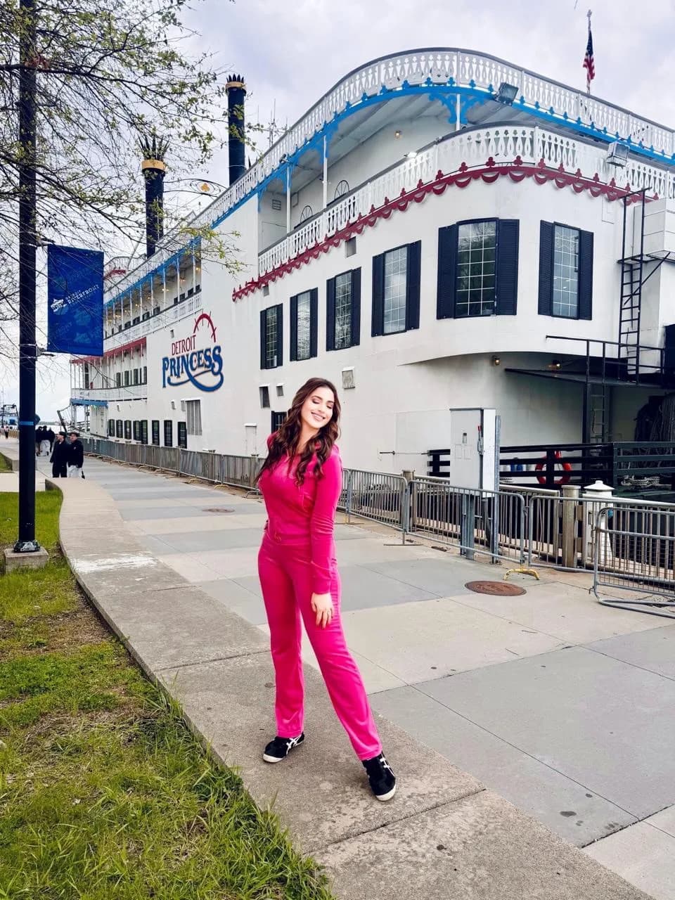 Samaira Sandhu smiling in a bright pink velour tracksuit and sneakers, standing on a concrete path next to the white "Detroit Princess" paddlewheel boat.