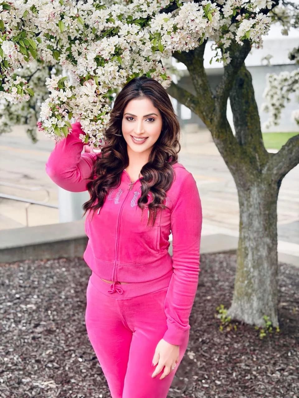 Samaira Sandhu, an Indian actress, smiling brightly in a vibrant pink velour tracksuit, standing next to a tree with white spring blossoms.