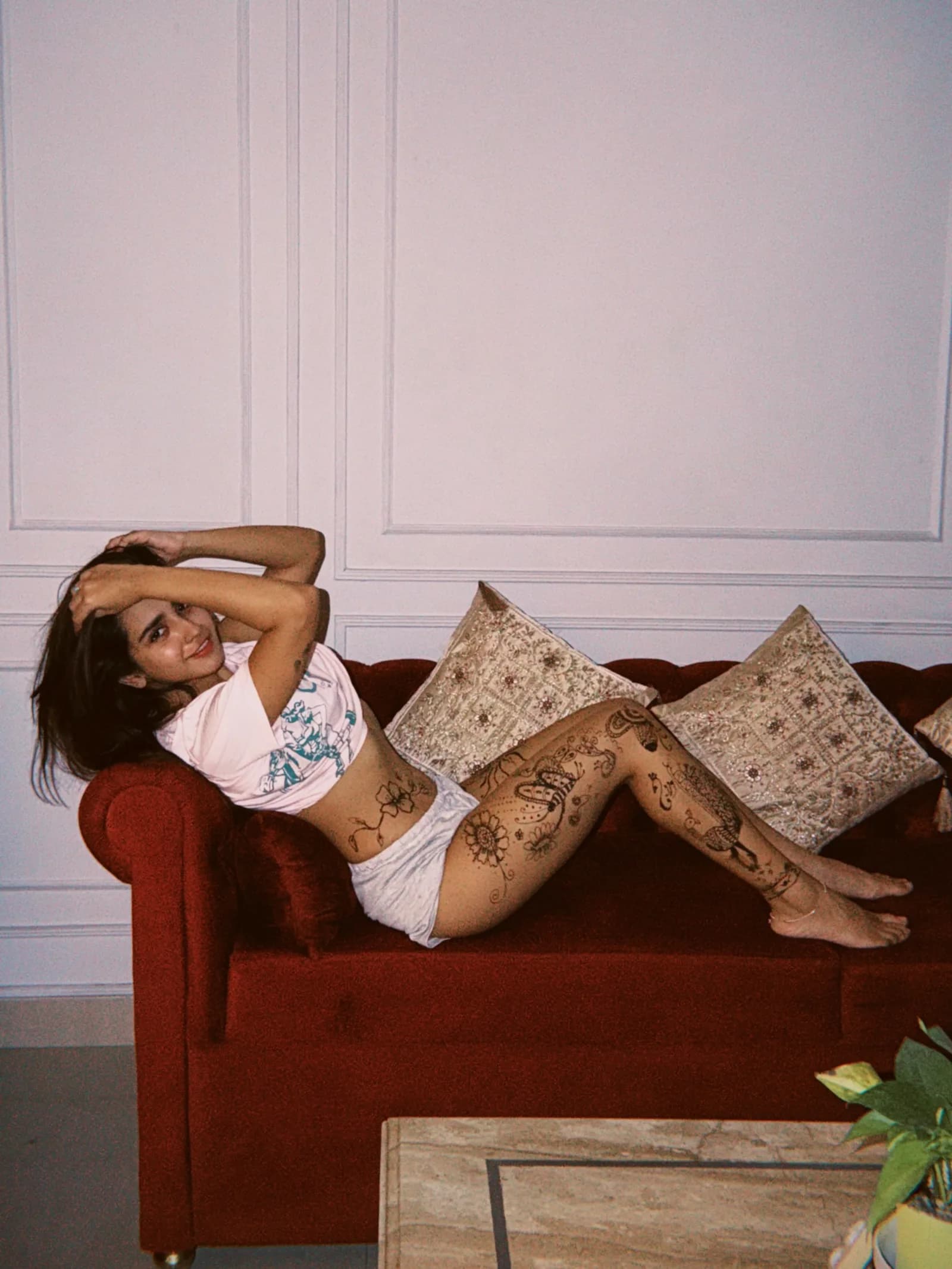 Roshni Walia reclining on a red velvet sofa, smiling at the camera, wearing a pink crop top and shorts, with intricate henna-style body art covering her legs and hip.