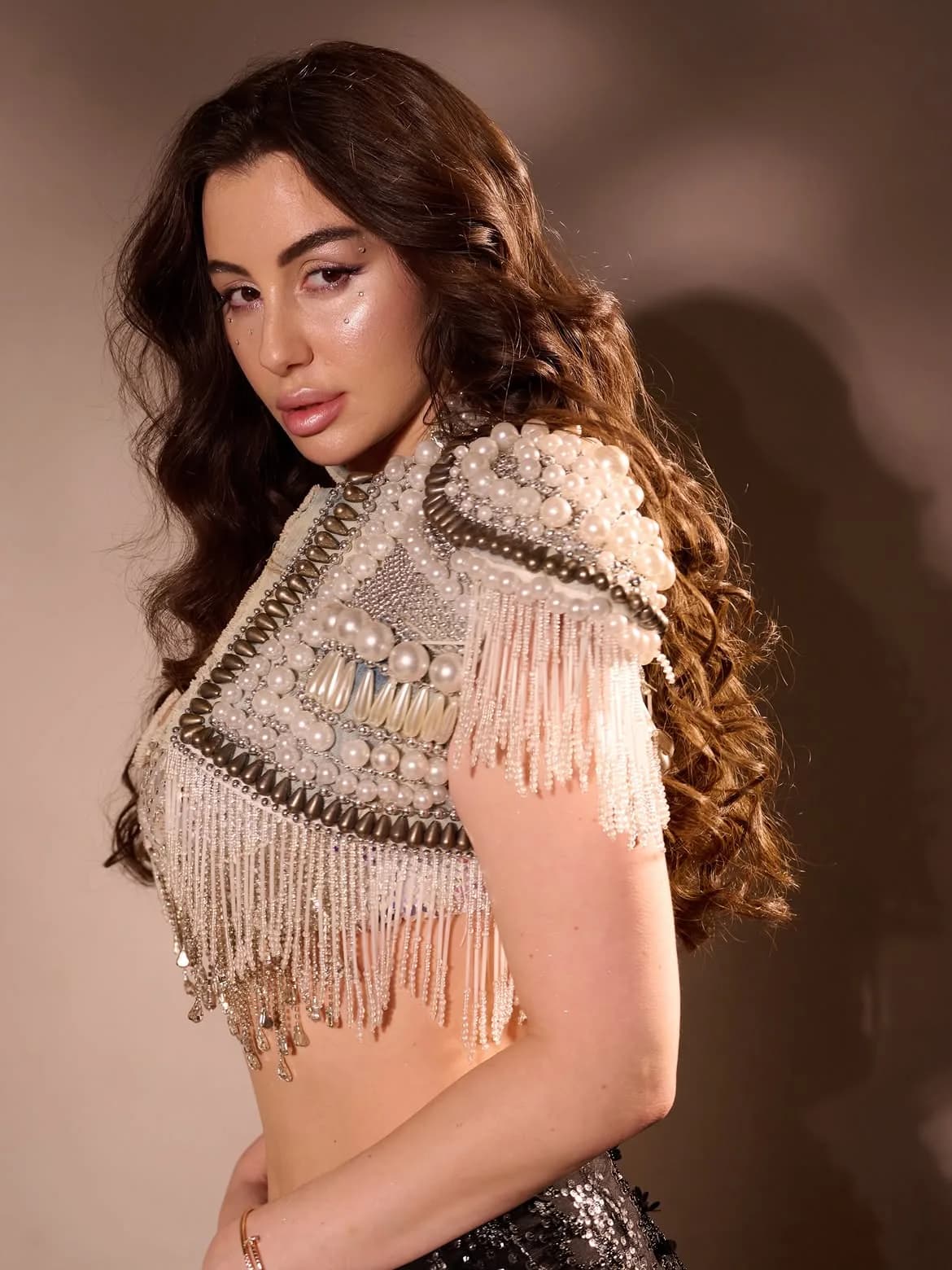 Giorgia Andriani posing in a white pearl and bead embellished crop top with fringe, featuring long wavy dark brown hair and glamorous makeup with eye glitter.
