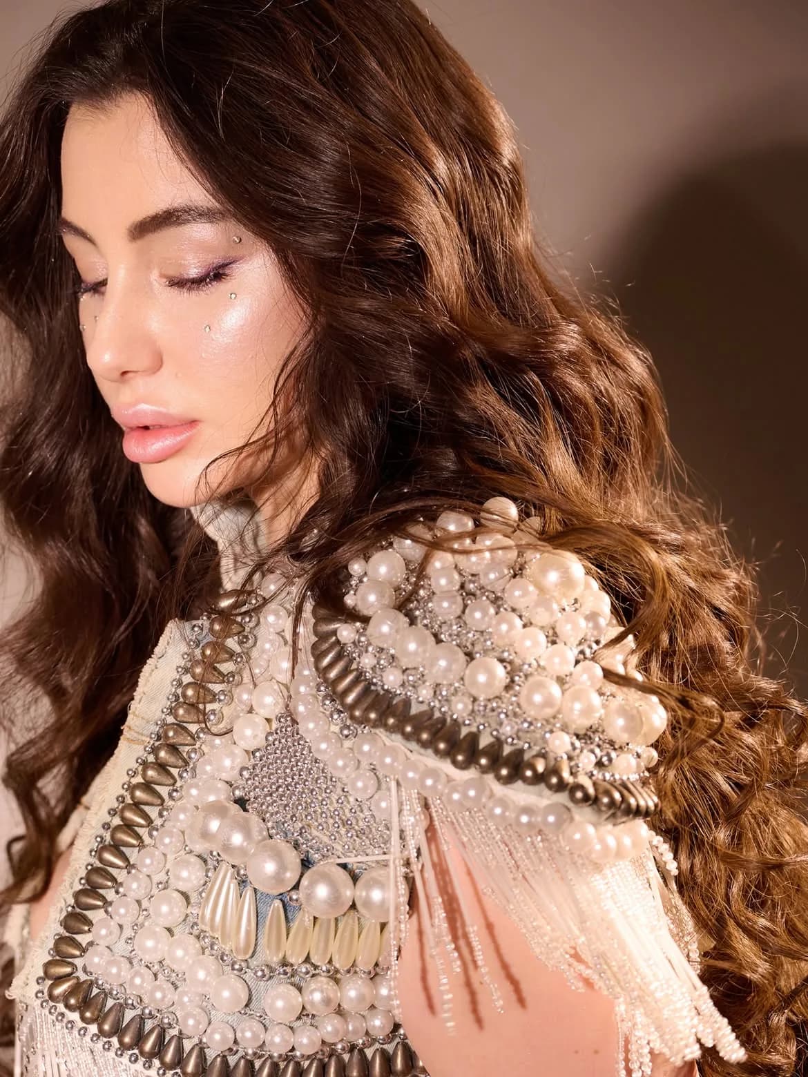 Close-up side profile of Giorgia Andriani with eyes closed, wearing a heavily pearl and stud-embellished white and bronze garment, with long wavy brown hair.