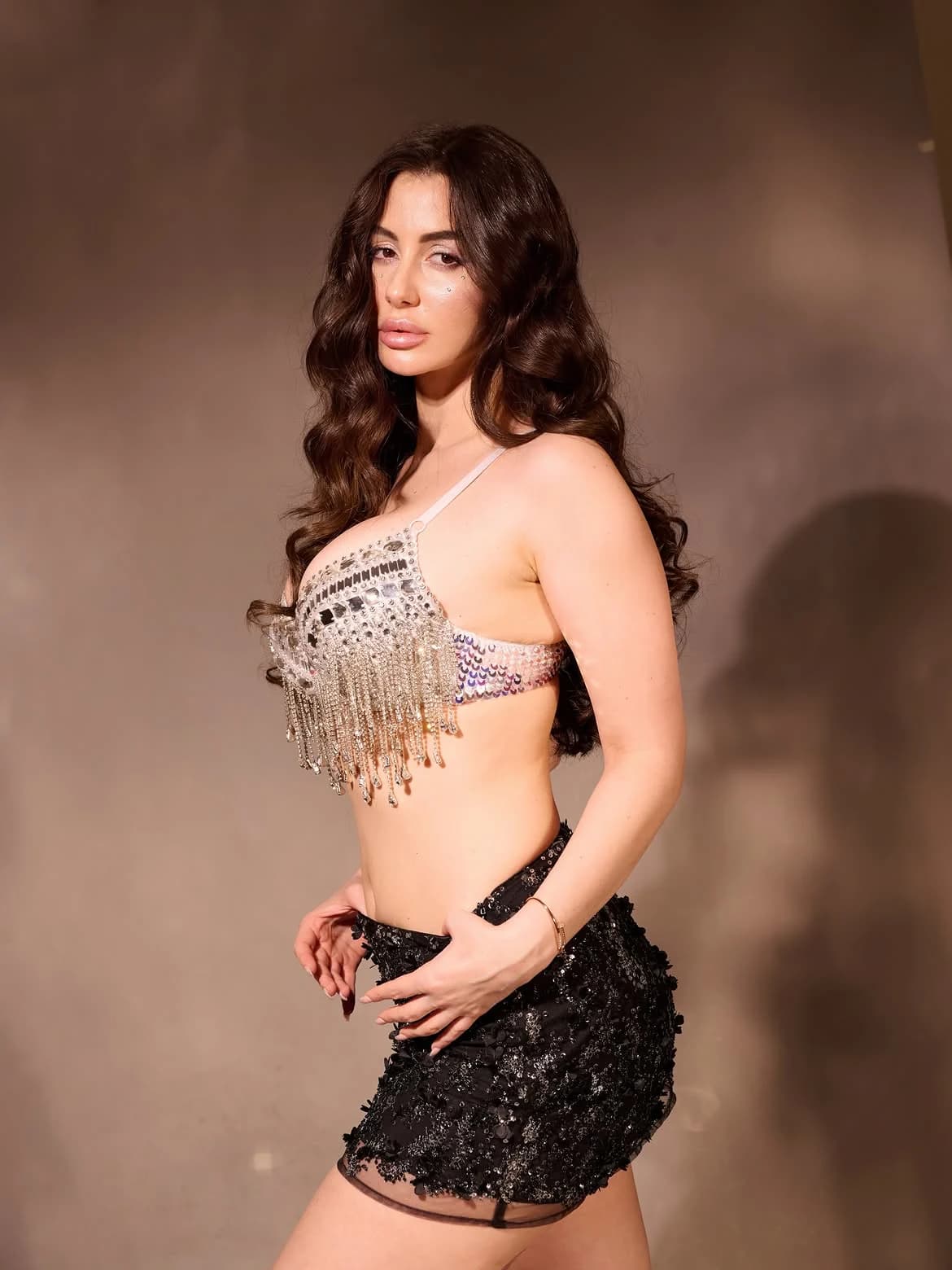 Giorgia Andriani posing in a shimmering silver embellished bra-top with fringe and a black sequined mini-skirt, against a brown background.