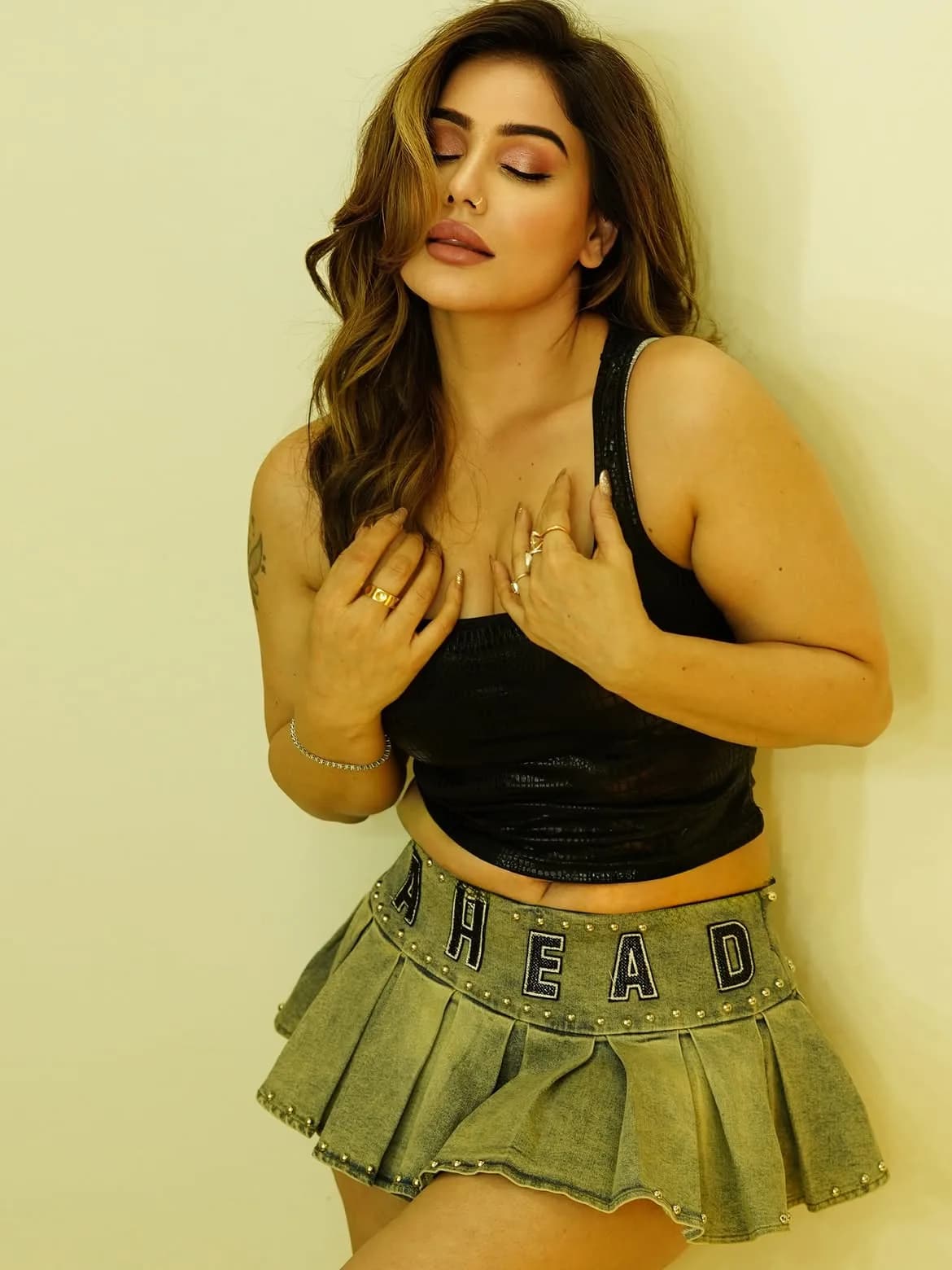 Kangna Sharma posing against a light wall, wearing a black sleeveless top and a studded denim mini skirt, with her eyes closed and hands near her chest.