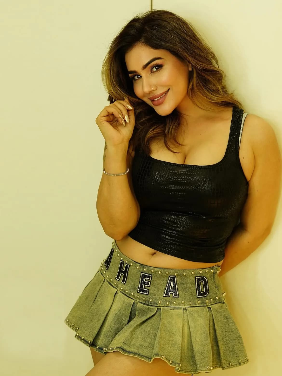 Kangna Sharma smiling, wearing a black textured crop top and a studded pleated denim mini skirt, leaning against a wall.