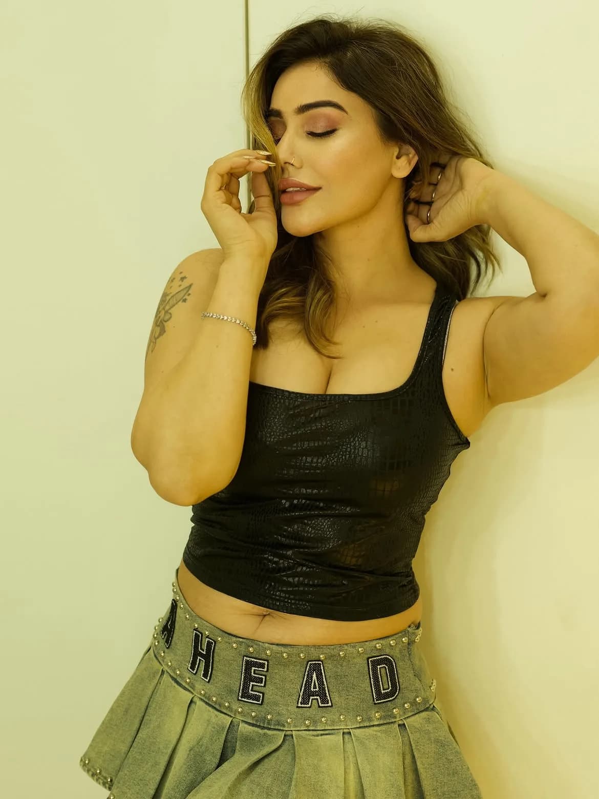 Kangna Sharma posing against a wall, wearing a black textured tank top and a studded denim mini-skirt, with her eyes closed and a hand near her face. Her midriff, cleavage, and armpits are visible.