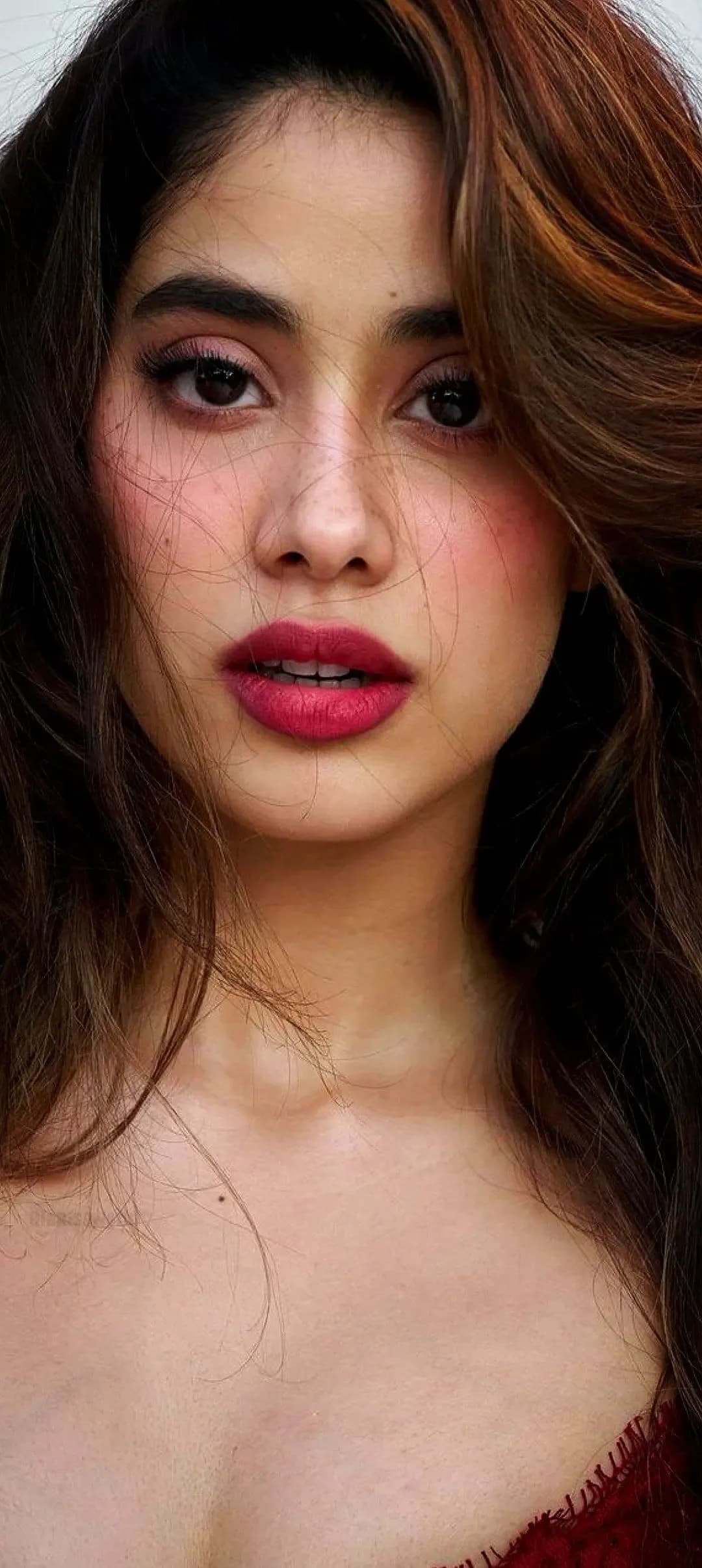 Close-up portrait of Janhvi Kapoor with long brown hair, bold red lipstick, and expressive eyes, wearing a red top.