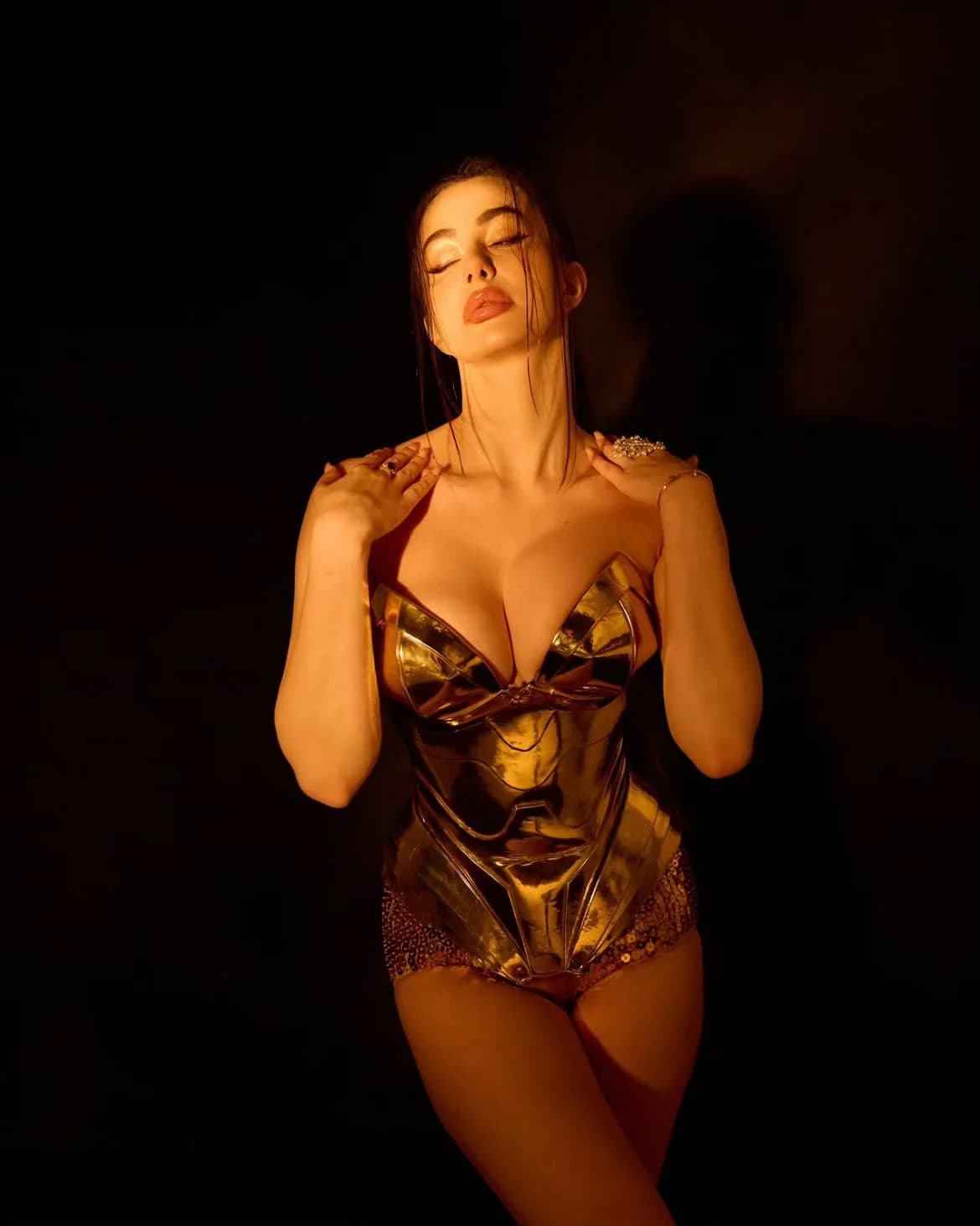 Giorgia Andriani in a golden metallic bodysuit, eyes closed, hands on shoulders, against a dark background.