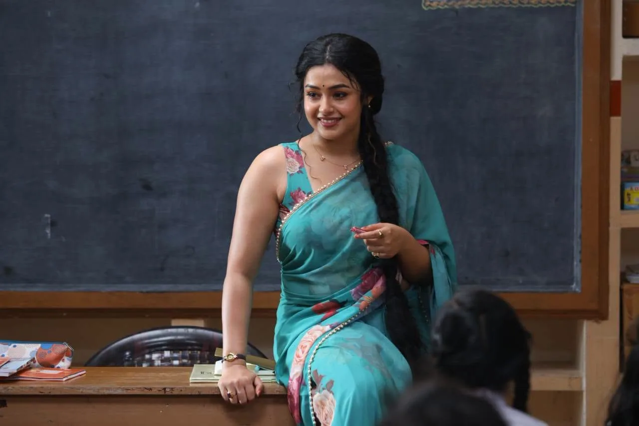 Mokksha as Meenu Miss in a teal floral saree sitting on a classroom desk