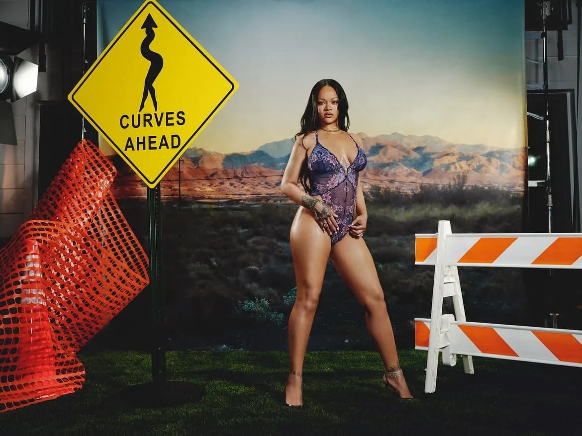 Rihanna wearing a purple sheer lace bodysuit next to a Curves Ahead sign.