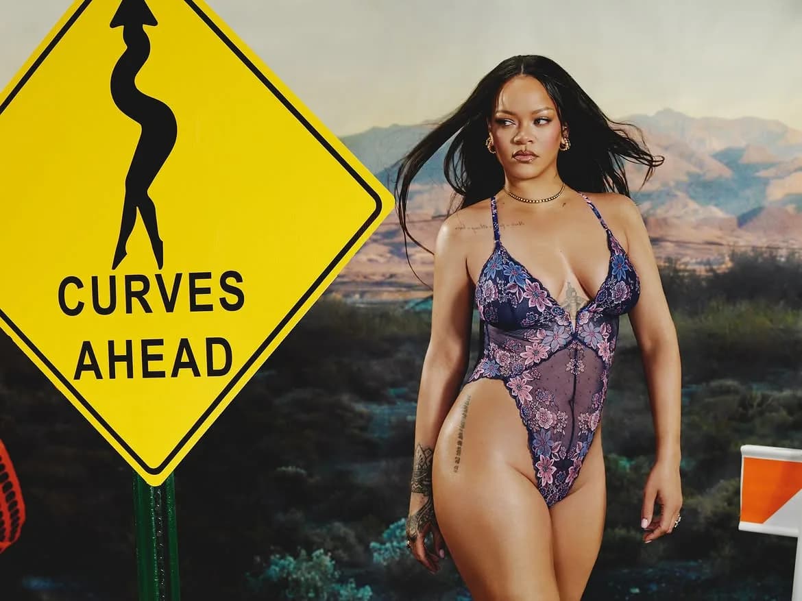 Rihanna wearing a sheer floral lace bodysuit posing beside a yellow Curves Ahead road sign in a desert.