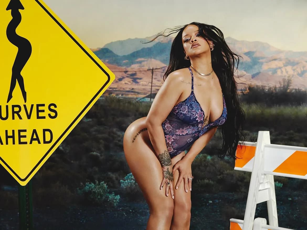 Rihanna posing in a blue floral lace bodysuit next to a yellow 'Curves Ahead' sign in a desert setting.