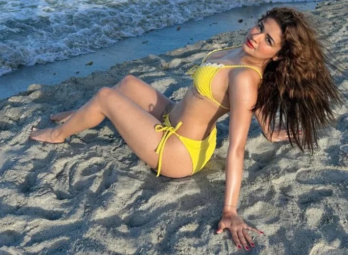 Avneet Kaur posing on a sandy beach wearing a yellow bikini with ocean waves behind her.